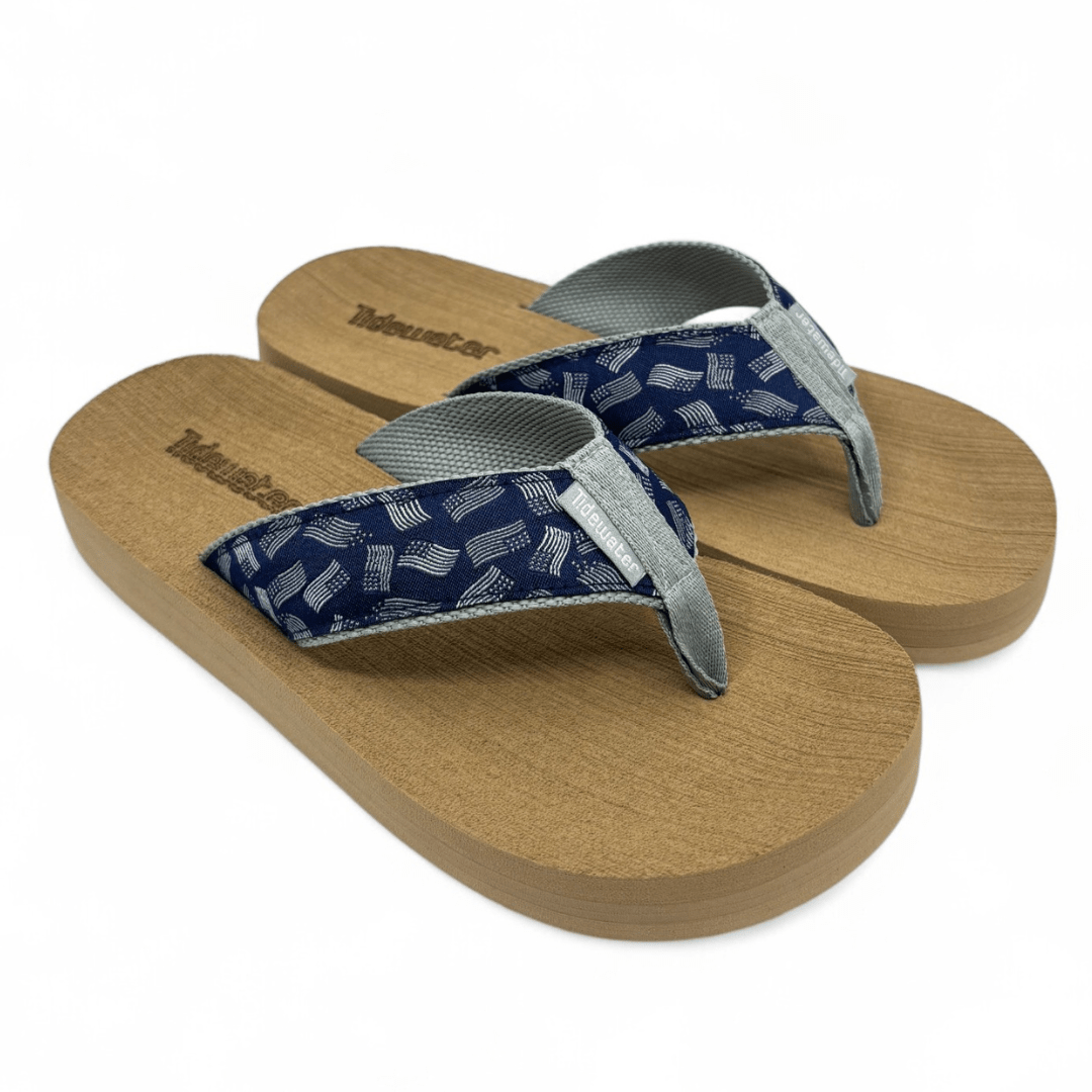 Pair of flip flops with tan comfort footbed and navy straps patterned with white American flags and gray woven lining

