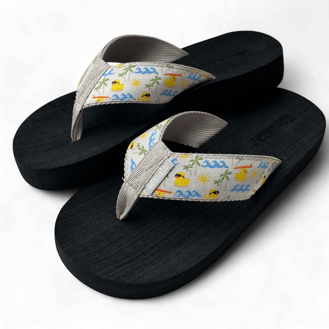 Pair of flip flops with black footbed and beige straps featuring rubber duck, wave, sun, and palm tree beach scene pattern

